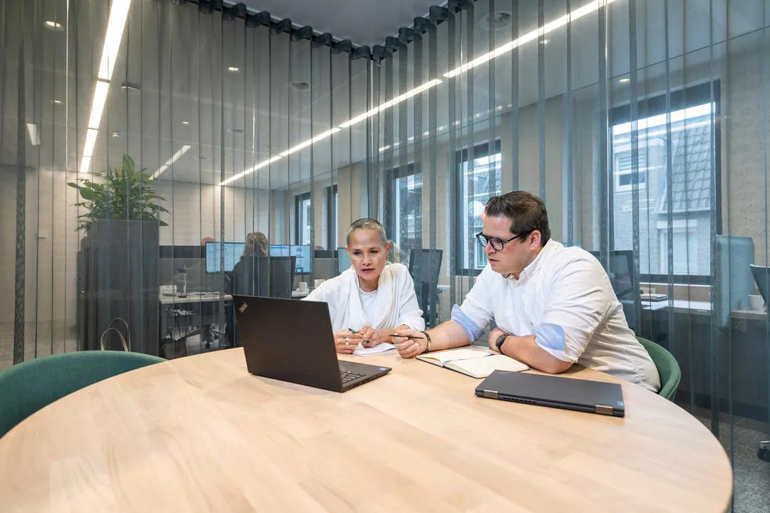 Twee medewerkers zitten in een vergadering te overleggen en aantekeningen te maken, kijkend naar het