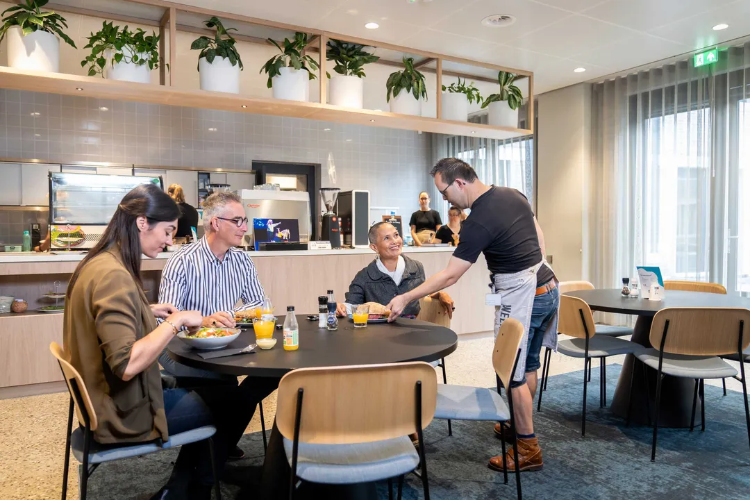 In het lunch- werkcafé zit een aantal collega’s aan tafel te lunchen. De ober serveert een gerecht. In het lunch- werkcafé zit een aantal collega’s aan tafel te lunchen. De ober serveert een gerecht.