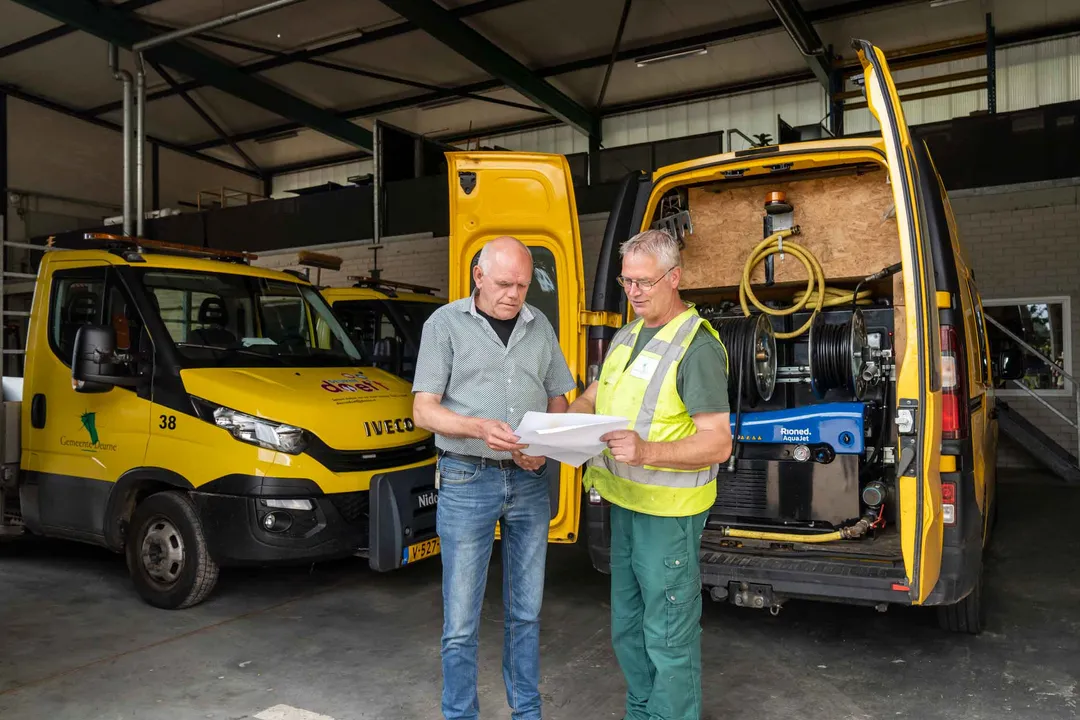 Twee buitendienst medewerkers bespreken samen wat er op het papier geschreven of afgebeeld staat.