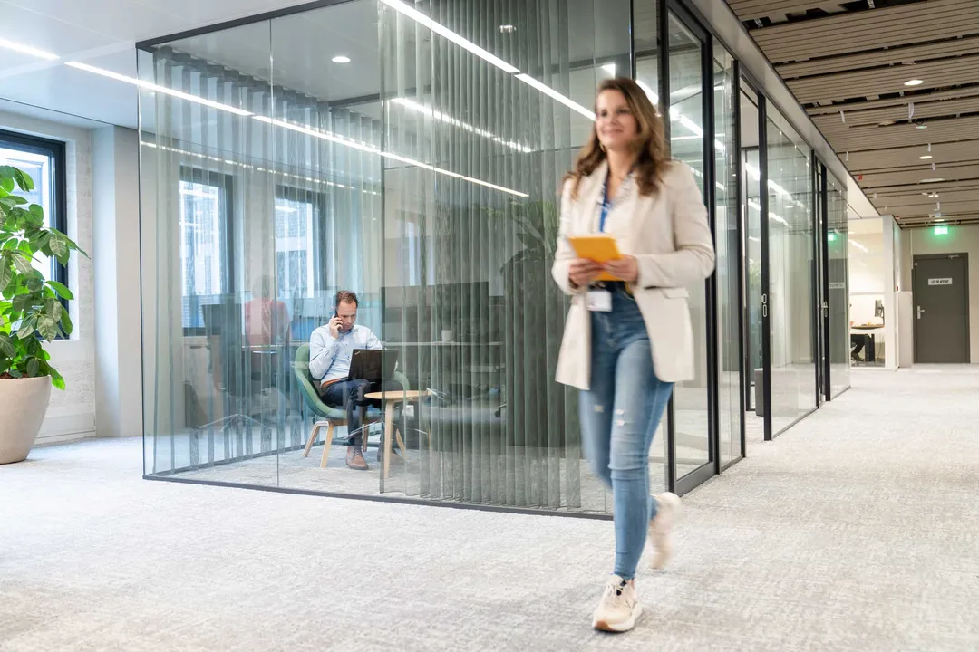 Een medewerker is in een gesloten ruimte aan het bellen. Er loopt een andere medewerker langs. Een medewerker is in een gesloten ruimte aan het bellen. Er loopt een andere medewerker langs.