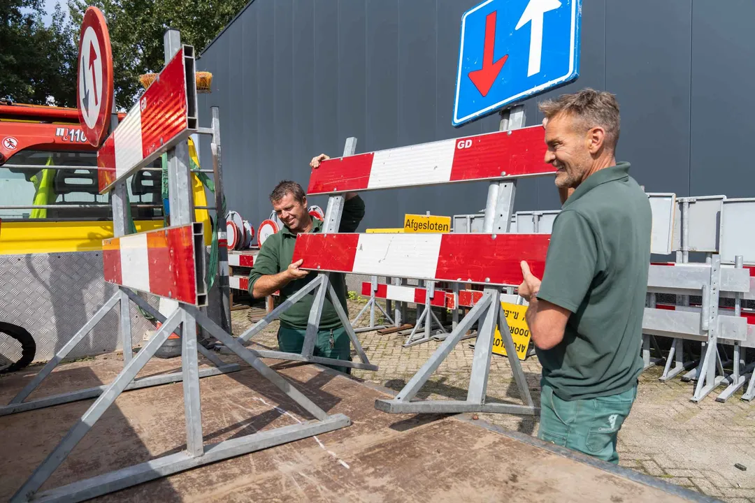Twee collega’s van de buitendienst tillen een omleidingsbord op de aanhangwagen. Twee collega’s van de buitendienst tillen een omleidingsbord op de aanhangwagen.
