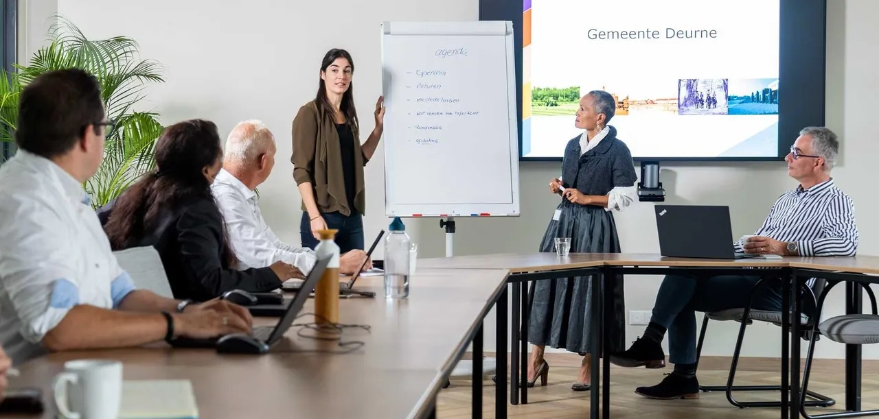 Een aantal medewerkers zit en staat in een vergadering en zij maken gebruik van het scherm.